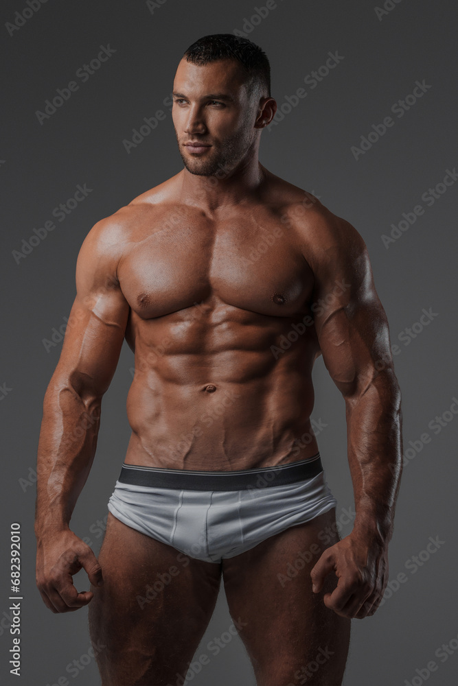 Portrait of a rugged man with a well-groomed model appearance, wearing white briefs, and showcasing his bare muscular torso against a gray background