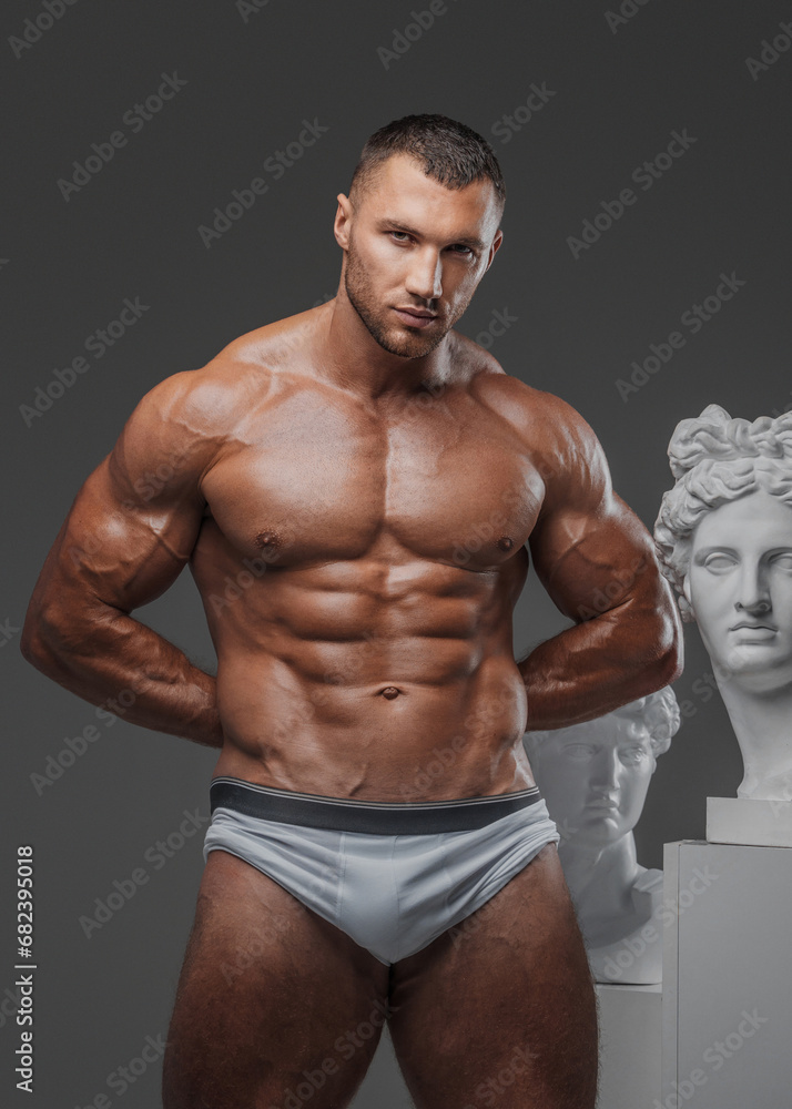 Fototapeta premium Handsome man exuding a rugged charm, with a flawless muscular chest, posing proudly beside ancient Greek statues against a gray backdrop