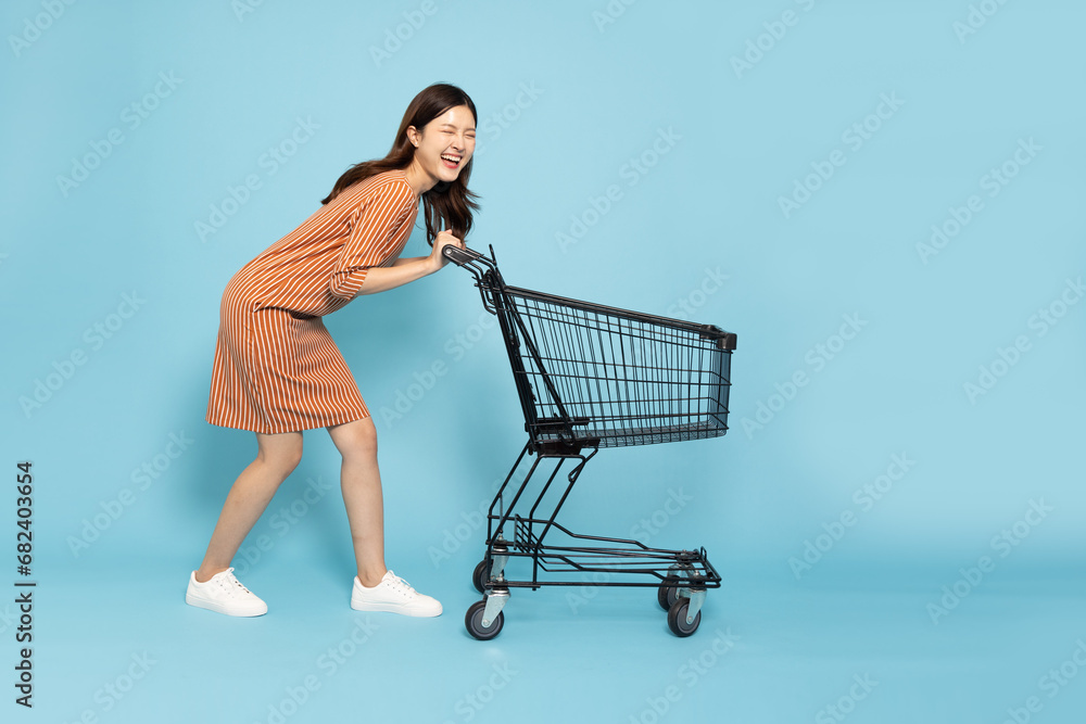 Full length portrait of young Asian woman pushing an empty shopping cart or shopping trolley isolated on blue sky background