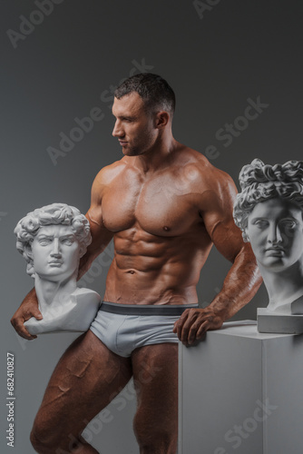 Handsome man exuding a rugged charm confidently showcasing his immaculate muscular physique while standing beside ancient Greek statues against a gray backdrop