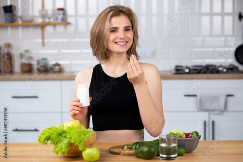 Beauty supplement. Attractive young woman holding vitamin capsule and glass of  water  at the kitchen. Healthy lifestyle, healthy diet nutrition concept photo