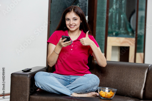 Indian young girl watching phone while sitting on sofa at home and showing thumbs up towards the mobile © Photographielove
