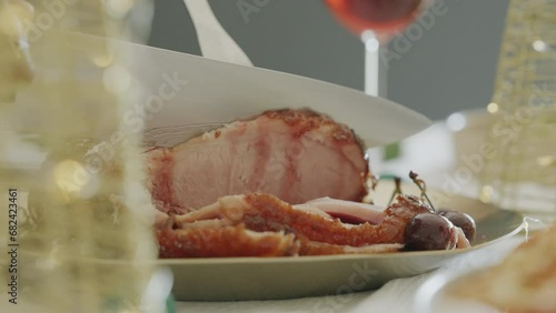 Woman slices Christmas ham meat thick pieces on a wooden table. Chef cooking Christmas meal dinner or lunch, smoked bacon in family home. Close up of festive decorated table. Holiday season.