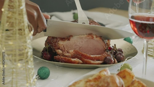 Woman slices Christmas ham meat thick pieces on a wooden table. Chef cooking Christmas meal dinner or lunch, smoked bacon in family home. Medium shot of festive decorated table. Holiday season.