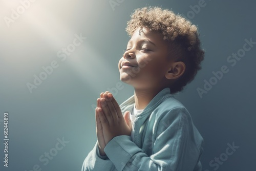 Little black boy on knees holding hands and praying in the morning, pastel neutral background. Christianity, faith, spirituality, religion, salvation, peace, faith concept. Kid praying to God © jchizhe