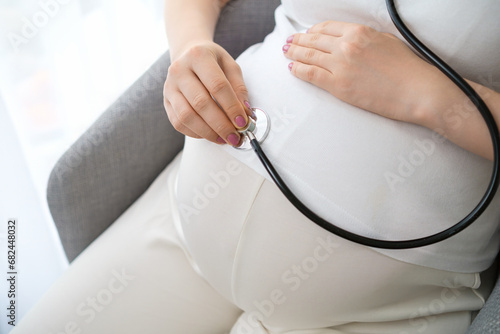 Pregnant woman, dressed in a light outfit listening to her stomach with a stethoscope. 