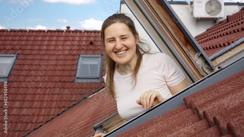 Happy smiling and laughing woman looking out of the open attic window in her apartment under the red tiled roof © Кирилл Рыжов