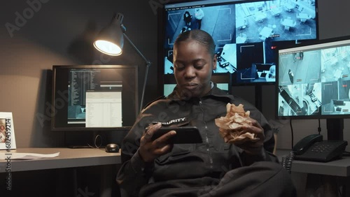 Medium shot of young careless African American security officer eating snack and watching video on phone not monitoring CCTV footage in guard room