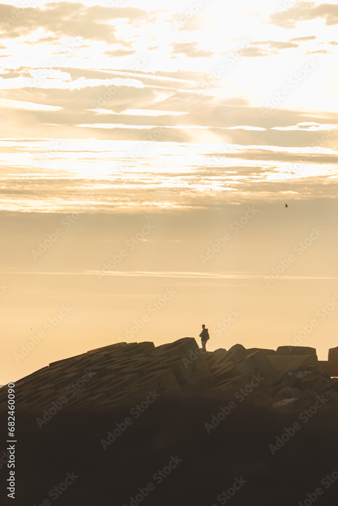 Man aged 25-29 standing on a seawall on the Atlantic Ocean in the yellow-orange rays of the sun near Oostende, Belgium