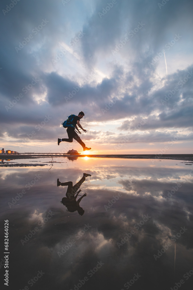 Fototapeta premium Brown-haired man with a backpack, aged 25-29, jumps into a pool of water on the beach during sunset. Catching the sun between the ground and his feet. Oostende, Belgium