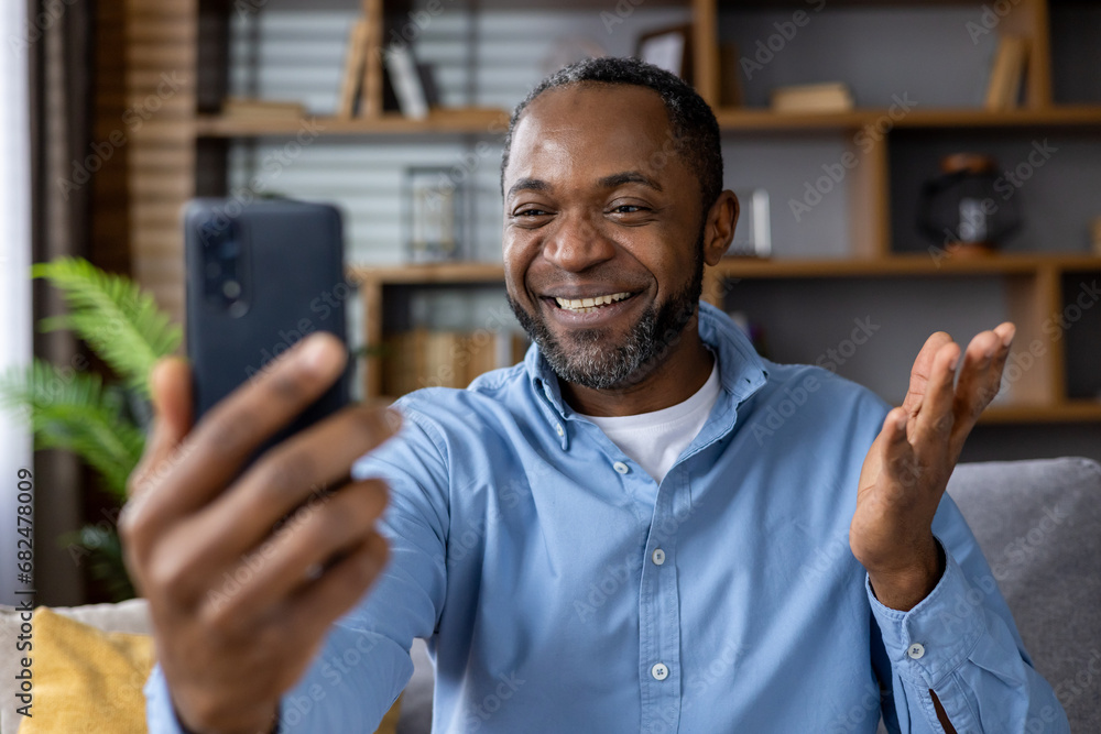 Senior man sitting on sofa in living room, African-American man holding phone in hands, chatting using video call app, smiling joyfully at interlocutor. Senior man sitting on sofa in living room, African-American man holding phone in hands, chatting using video call app, smiling joyfully at interlocutor.