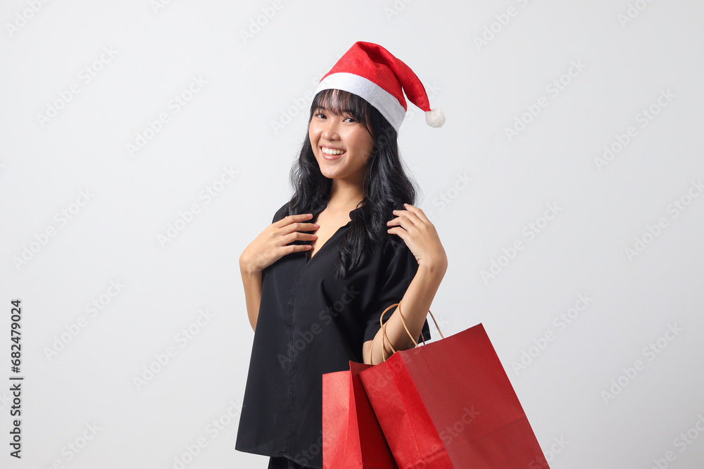 Portrait of attractive Asian woman with red Santa hat feeling happy while purchasing stuff. Excited girl holding shopping bag. New year and christmas concept. Isolated image on white background Portrait of attractive Asian woman with red Santa hat feeling happy while purchasing stuff. Excited girl holding shopping bag. New year and christmas concept. Isolated image on white background