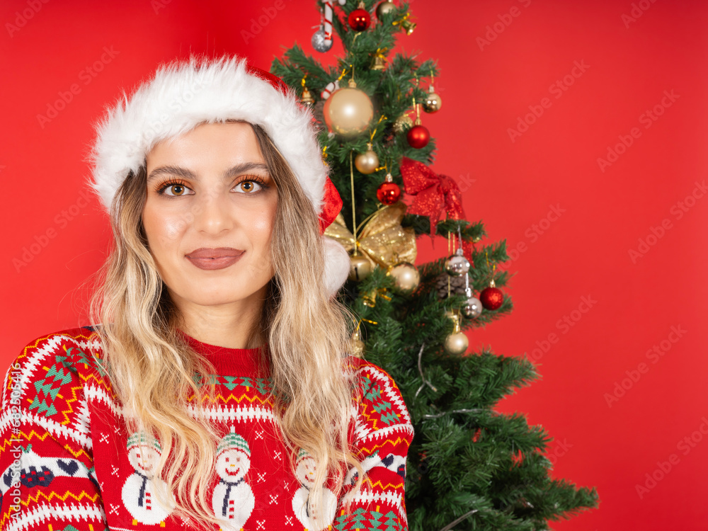 Winter holiday concept, portrait of young beautiful caucasian woman posing winter holiday concept. Standing near Christmas tree. Red studio background. Wear santa hat, knitted sweater. Copy space.