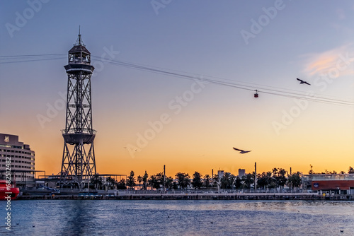 Barcelona, Spain - November 26, 2021: Steel Torre Jaume I tower and Port Vell Aerial Tramway cable car against purple-orange sky at dusk in Barcelona. Cityscape and seascape of Port Vell at sunset photo