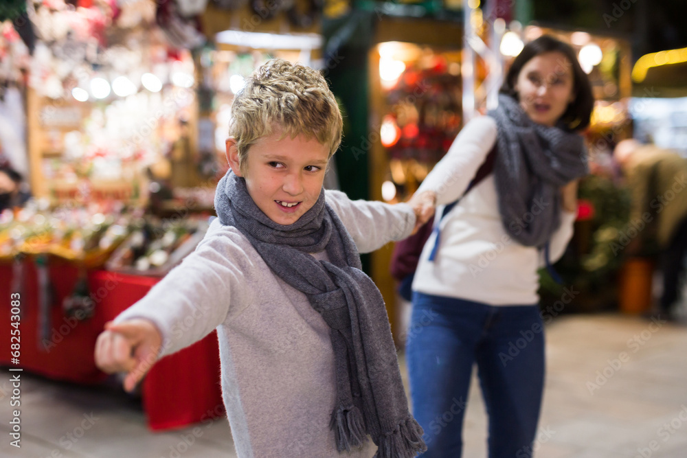 Portrait of tween boy pointing to desired thing and pulling mothers hand during walk at festive fair on Christmas Eve