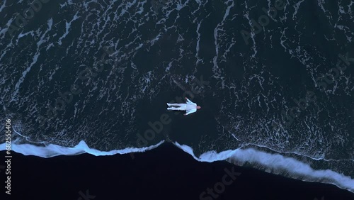 Dramatic view of desperate male wearing white clothes lying motionless on black sanded beach while wild foaming waves strike the shore, washing the man. High quality 4k footage