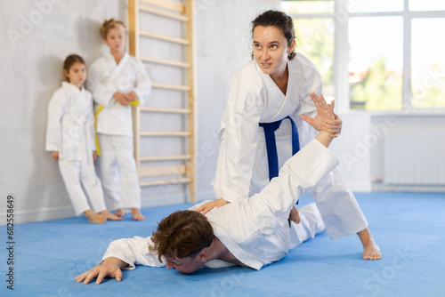 Concentrated young woman attending family karate class, applying armlock technique to husband while preteen children in kimonos watching training sparring in background