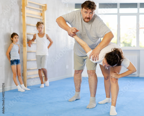 Father and mother demonstrating self-defense techniques to preteen children standing against wall in gym and watching training bout between parents practicing painful armlock..