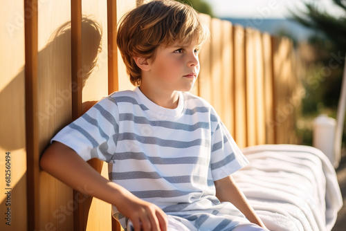 Calm boy in a striped T-shirt sitting near a fence while looking away during summer holiday at a sunny resort.