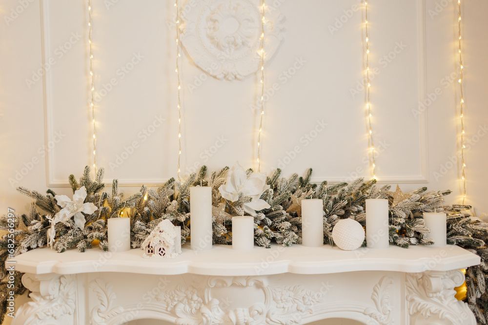 Luxury white fireplace with Christmas decoration, fir branch, candles, xmas glittering baubles, shiny flowers ornaments. Festive wall with illuminate garland warm lights . New year 2024 holidays.
