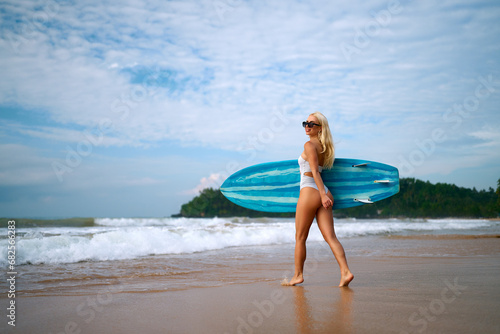 Blonde woman in white bikini carries blue surfboard on sandy beach, prepares for surfing session in ocean waves, embodies active summer vacation and water sports lifestyle.