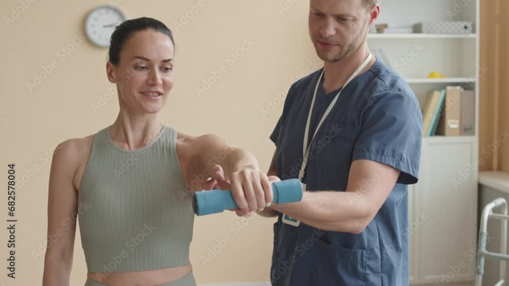 Male physiotherapist helping female patient with doing dumbbell front raise during rehabilitation therapy in clinic Male physiotherapist helping female patient with doing dumbbell front raise during rehabilitation therapy in clinic
