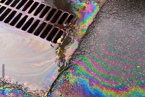 An oil slick against the backdrop of an asphalt road flows into a storm drain through a grate.