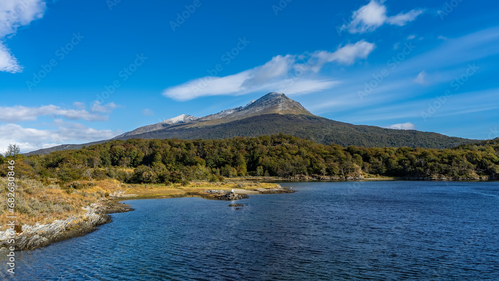 Beautiful blue lake. Yellowed grass and thickets of trees grow on the shore. A picturesque mountain against a blue sky and clouds. Argentina. Lapataia Bay. Tierra del Fuego National Park. Ushuaia