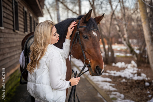 Young beautiful rider woman blonde with long hair in white clothes standing near horse