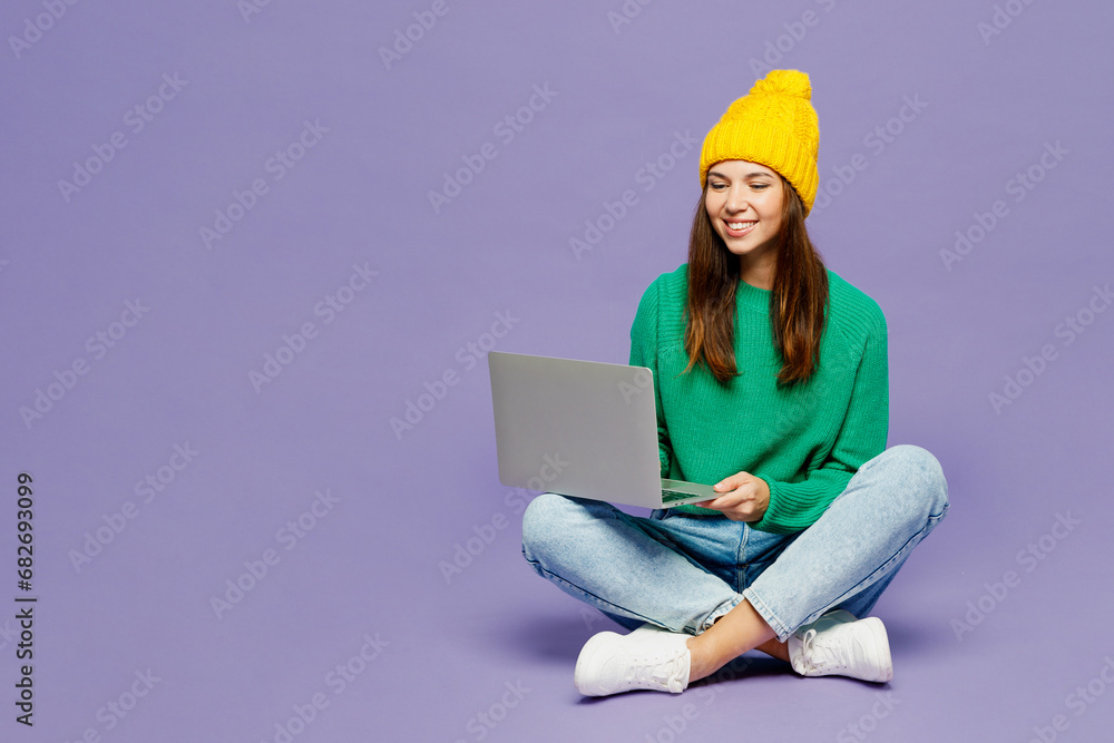 Full body young IT woman she wear green sweater yellow hat casual clothes sit hold use work on laptop pc computer isolated on plain pastel light purple background studio portrait. Lifestyle concept.