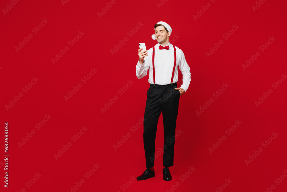 Full body merry cool young man wear white shirt Santa hat hold use mobile cell phone type message posing isolated on plain red background studio Happy New Year Christmas celebration holiday concept Full body merry cool young man wear white shirt Santa hat hold use mobile cell phone type message posing isolated on plain red background studio Happy New Year Christmas celebration holiday concept