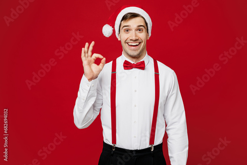 Merry young smiling satisfied man wearing white shirt Santa hat posing look camera show ok okay gesture isolated on plain red background studio. Happy New Year Christmas celebration holiday concept.