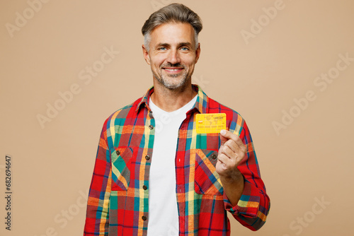 Adult cheerful smiling man wear red shirt white t-shirt casual clothes hold in hand mock up of credit bank card isolated on plain pastel light beige color background studio portrait Lifestyle concept