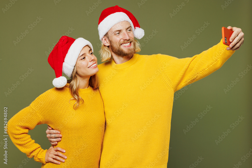 Merry young couple two friends man woman wear sweater Santa hat posing doing selfie shot on mobile cell phone isolated on plain green background. Happy New Year celebration Christmas holiday concept. Merry young couple two friends man woman wear sweater Santa hat posing doing selfie shot on mobile cell phone isolated on plain green background. Happy New Year celebration Christmas holiday concept.