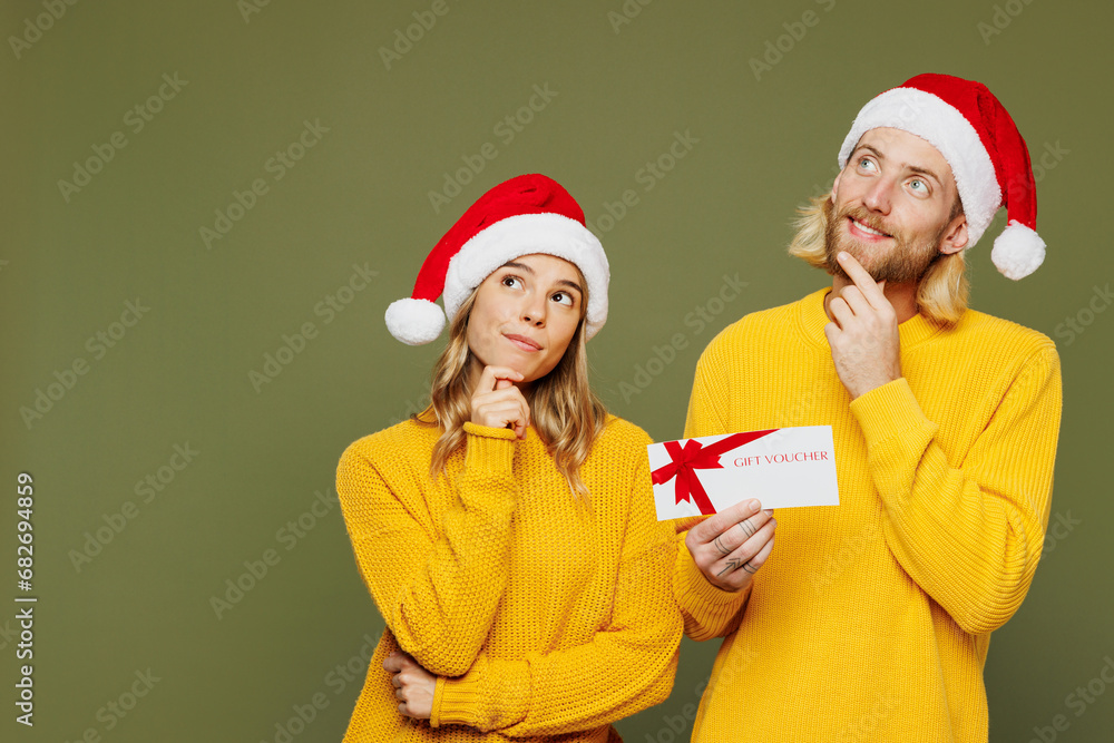 Merry minded young couple two friends man woman wear sweater Santa hat posing hold gift certificate coupon voucher card for store isolated on plain green background. Happy New Year Christmas concept. Merry minded young couple two friends man woman wear sweater Santa hat posing hold gift certificate coupon voucher card for store isolated on plain green background. Happy New Year Christmas concept.