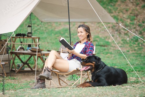 woman and a rottweiler dog on a camping trip in nature. relaxing woman read a book sitting on camping chair. a pet on vacation with his family, traveling with a dog, rest from the hustle and bustle. © nareekarn