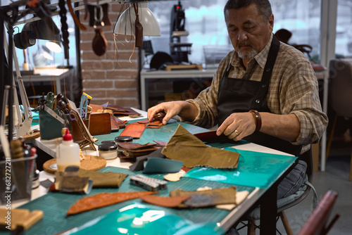 talented old serious man wearing apron and checked shirt using an awl makes holes for hand-stitching a leather product. closeup portrait.