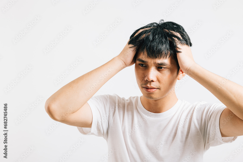 Portrait of a young Asian man with a sad and tired face, holding his head in despair due to stress and a severe headache. Studio shot isolated on white, symbolizing his distress. Portrait of a young Asian man with a sad and tired face, holding his head in despair due to stress and a severe headache. Studio shot isolated on white, symbolizing his distress.