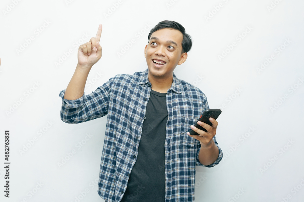 Naklejka premium Portrait Indonesian Asian young man smiling while looking at smartphone and index finger pointing up. Isolated white background