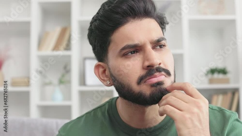 Thoughtful bearded man making important decision while solving life problem alone at home. Indian guy wearing casual green t-shirt sitting on grey couch in doubt. photo