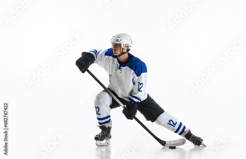 Young man, athlete, hockey player in uniform and helmet, standing on rink with stick against white studio background