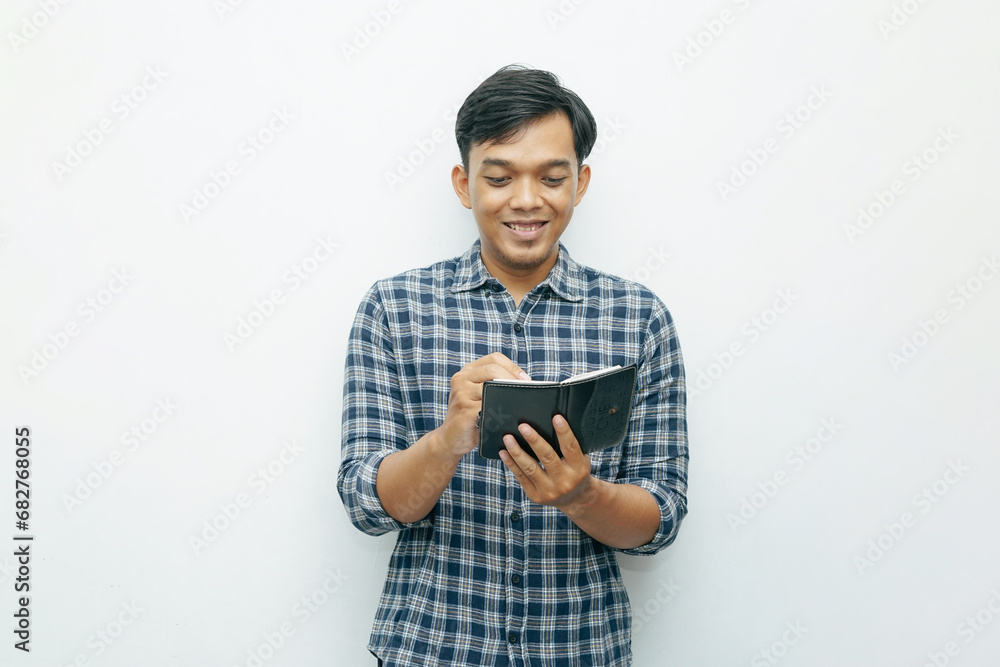 Portrait of young Asian Indonesian man smiling when writing on small notebook with ballpoint pen with isolated white background Portrait of young Asian Indonesian man smiling when writing on small notebook with ballpoint pen with isolated white background