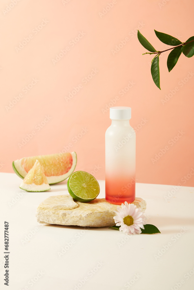An unlabeled bottle and half a lemon on a stone platform. Pomelo and flowers decorate the table with a pink background. Ideal space for advertising.