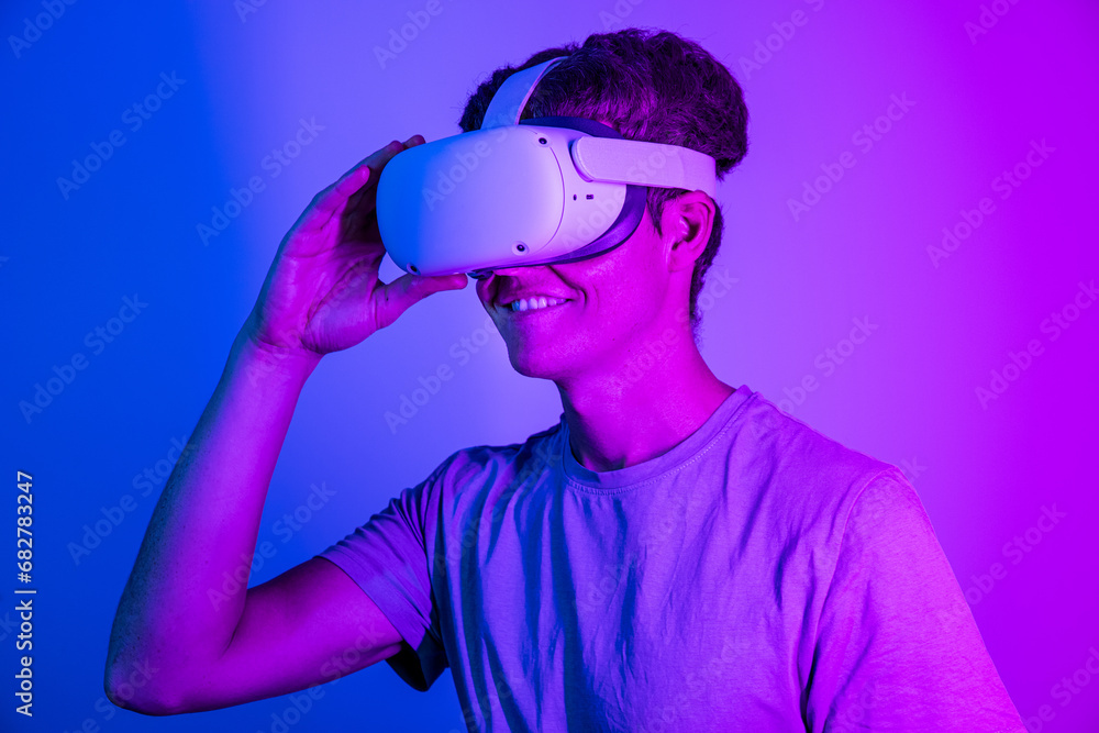 Smiling young guy with VR headset exploring virtual reality isolated on blue and pink neon background Smiling young guy with VR headset exploring virtual reality isolated on blue and pink neon background