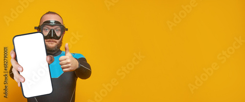 Scuba diver diving man holding smartphone with empty white screen display for new Application and showing thumb up on bright yellow studio wall background. Mockup banner