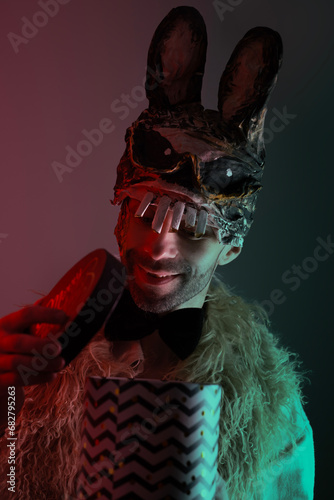 creepy hare man, young guy in cringe in scary bunny mask, holding box with gift, concept of Halloween party, dark zombie masquerade, theme of fear and horror photo