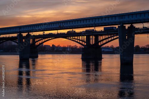 Beautiful view of the bridge over which cars drive at sunset. A river flows under the bridge, reflecting the sunset rays. Bridge in the city of Novosibirsk, Russia
