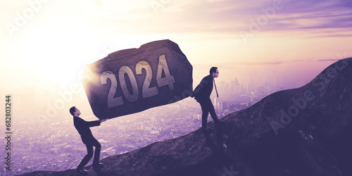 Two young businessmen lifting a stone with 2024 number while climbing on the cliff photo