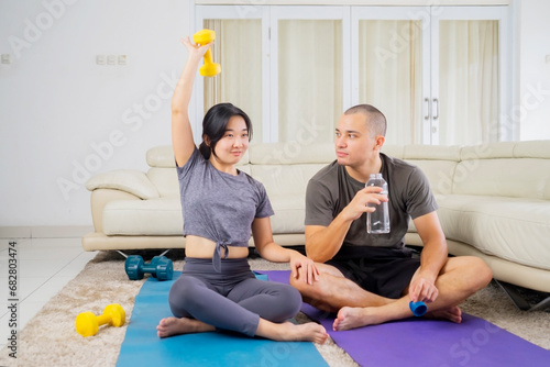 Young man drinking water while sitting with his exercising wife in the living room at home