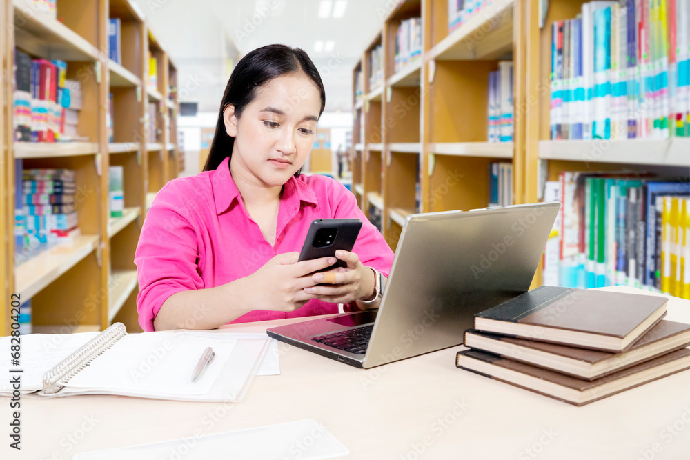 Female student using mobile phone while studying with a laptop computer and books in a library Female student using mobile phone while studying with a laptop computer and books in a library